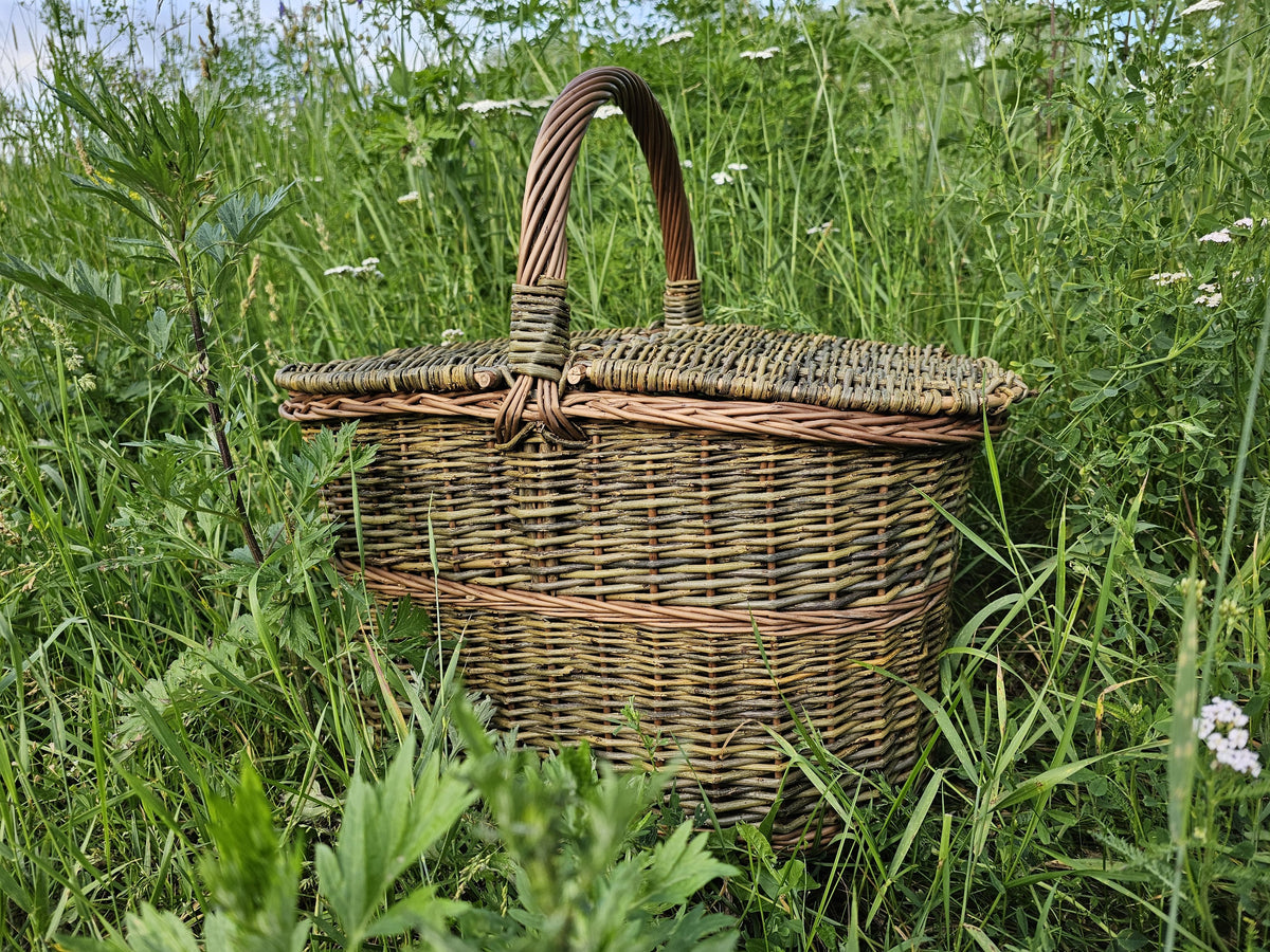 Pikniko pintinė su dangčiais ir lanko formos rankena pievoje, matyti pynimas ir tvirtas dugnas
