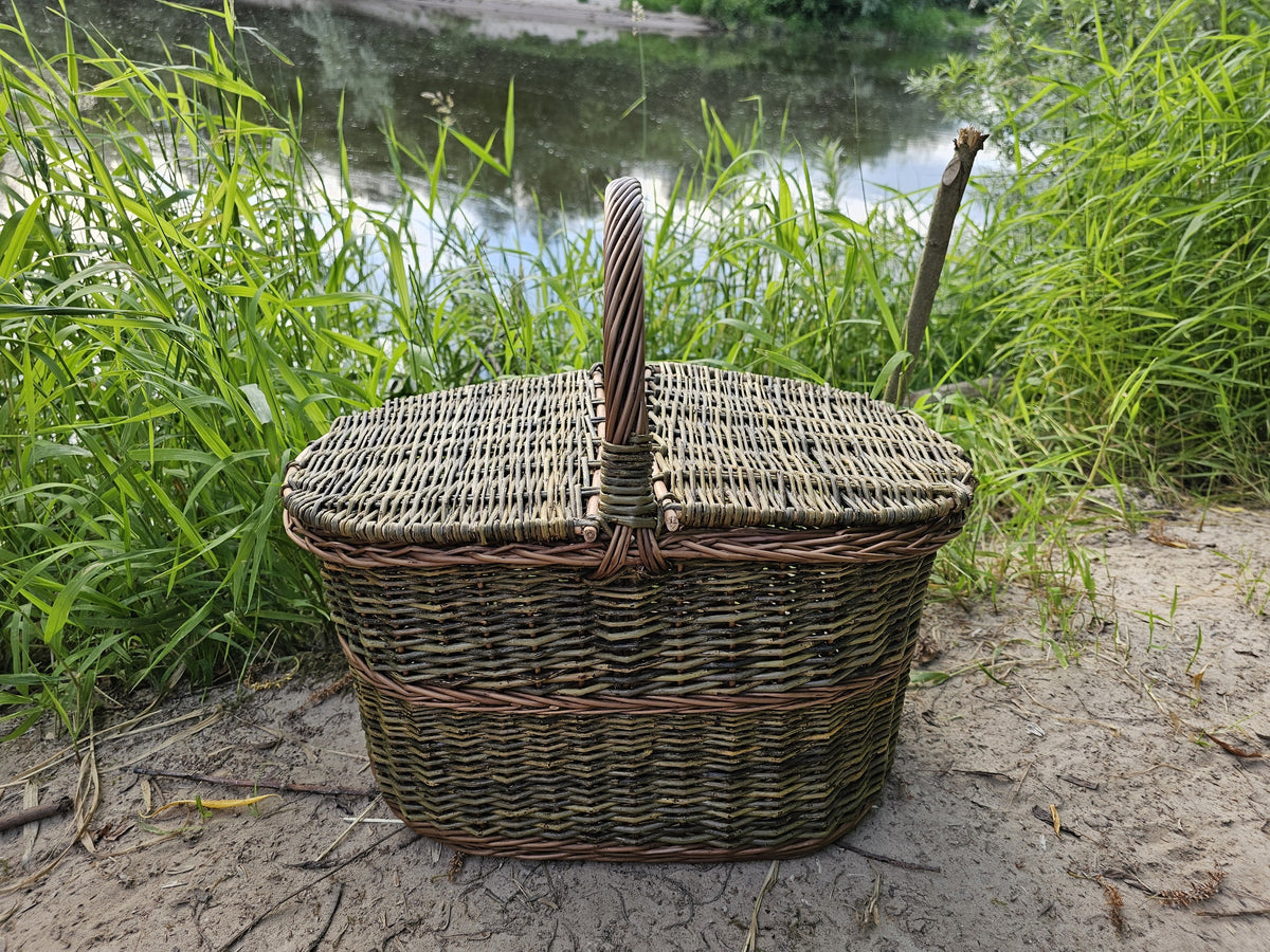 Ovali pikniko pintinė su dangčiu ir rankena per vidurį ant smėlio prie ežero tarp aukštos žolės