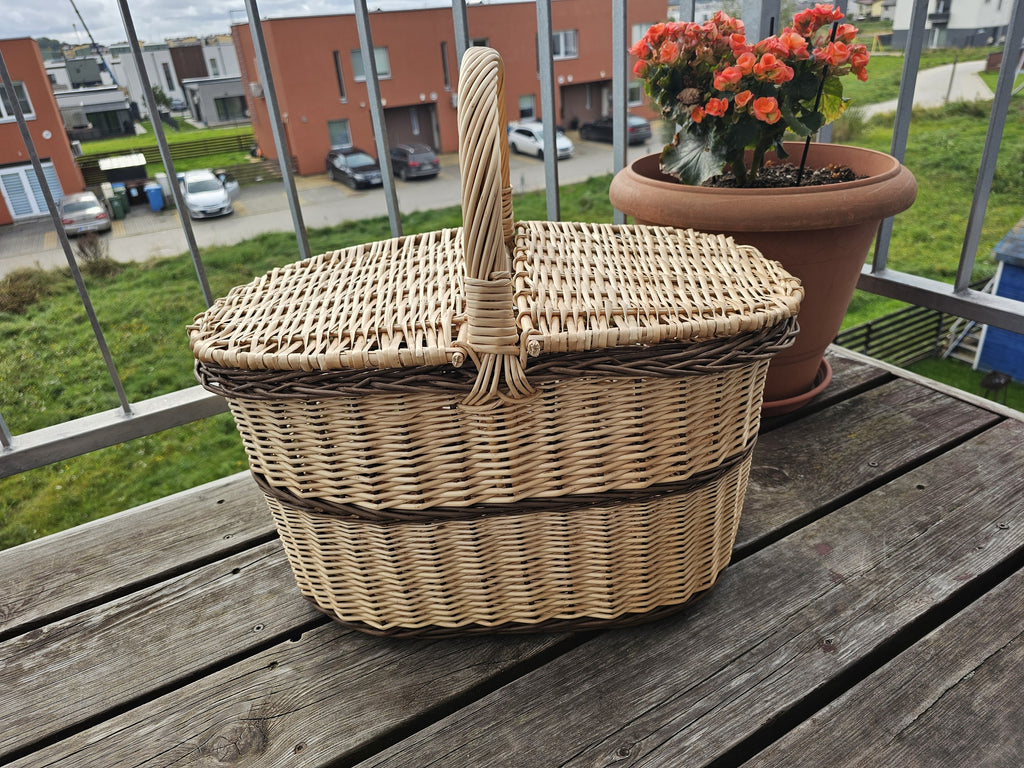 Ovali pikniko pintinė su dviem dangčiais ir rankena per vidurį ant medinio balkono stalo šalia gėlių vazono