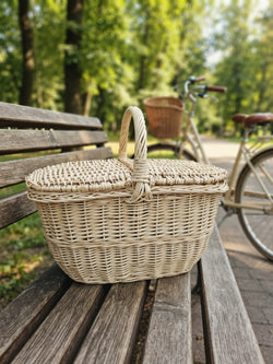 Small wicker picnic basket with a double lid and central handle, handwoven from willow, sitting on a park bench with a bicycle behind it.