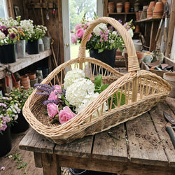 Großer Blumenkorb aus Weide mit hohem Henkel, gefüllt mit Rosen, Hortensien und Lavendel, auf einem Pflanztisch im Gartenhaus