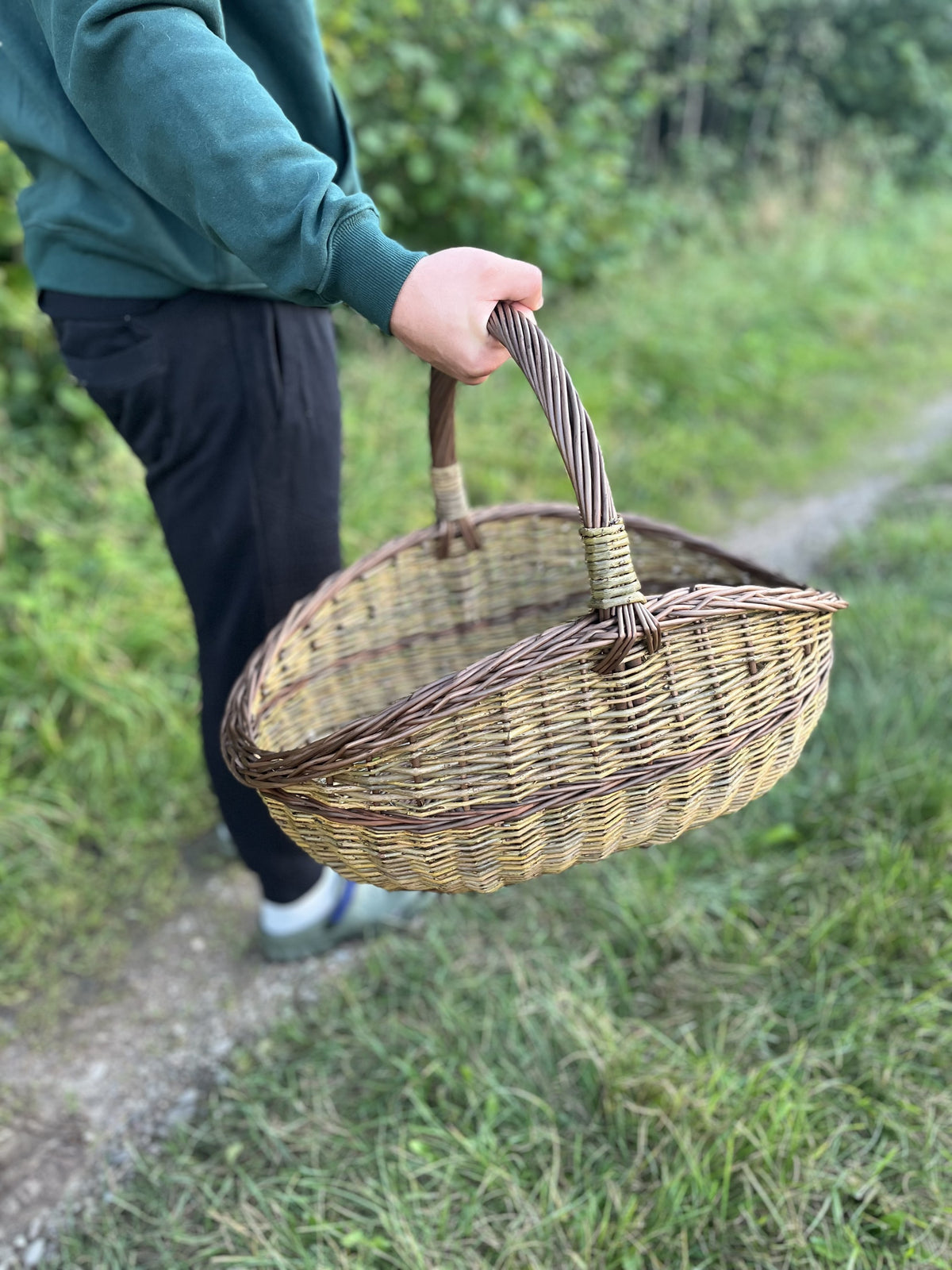 Žmogus neša pintinę viena ranka eidamas pakelės žole – matyti jos dydis