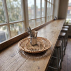 Ažūrinis vytelių padėklas su metaliniu puodeliu stalo įrankiams ant ilgo medinio kavinės baro prie aukštų langų