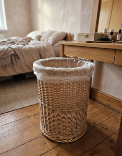 Round wicker waste basket with a bin liner, handwoven from willow, standing on a wooden floor beside a dressing table in a bedroom.
