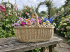 Shallow wicker harvest trug with a twisted handle full of roses, daisies and delphiniums on a garden bench among flower beds