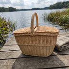 Uždaryta ovali iškylų pintinė su dvigubu dangčiu ir lanko formos rankena ant medinio lieptelio prie ežero, šalia vilnonis pledas