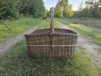 Large firewood basket with a tall twisted handle standing in the middle of a grassy farm track at sunset