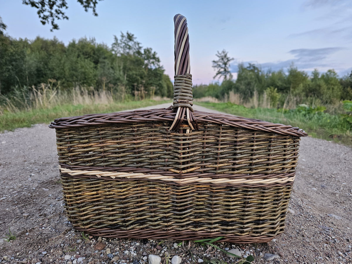 Side view of the large willow firewood basket with its tall upright handle on a gravel road under an evening sky