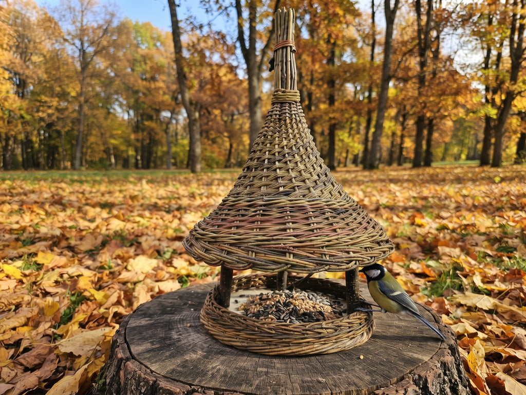 Aukšta vytelių lesykla su varpo formos stogeliu ant kelmo tarp rudens lapų, prie padėklo didžioji zylė