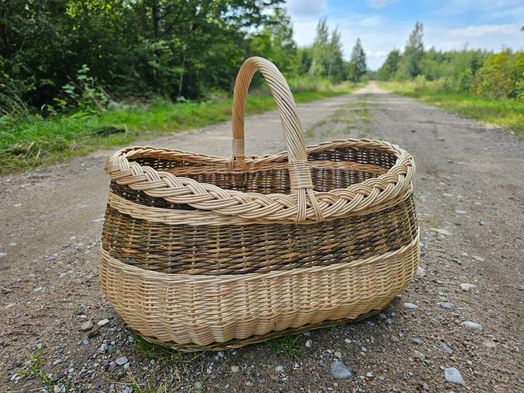 Didelė ovali pirkinių pintinė su aukšta rankena ir kasele pintu kraštu ant žvyruoto kaimo kelio tarp medžių