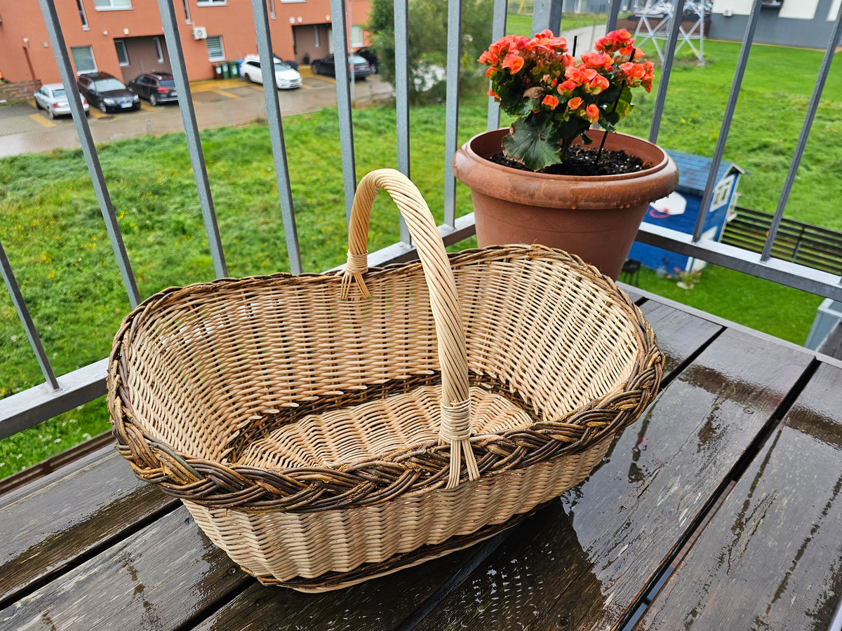 Didelė ovali pirkinių pintinė su kasele pintu kraštu ir aukšta rankena ant balkono stalo šalia gėlių vazono