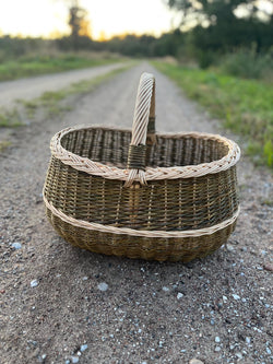 Large handwoven willow market basket with braided rim and arched handle standing on a gravel country road at dusk.