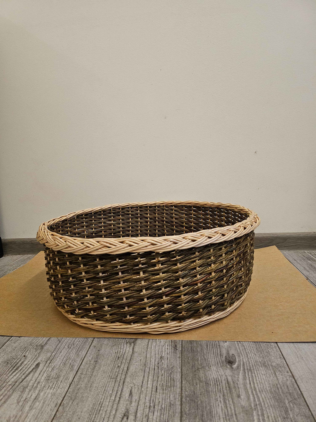 Apvali pintinė žemais šonais ir storu kasele pintu kraštu ant rudo popieriaus prie šviesios sienos