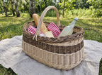 Large oval willow picnic basket with a baguette, checked napkins and a bottle of lemonade on a linen cloth