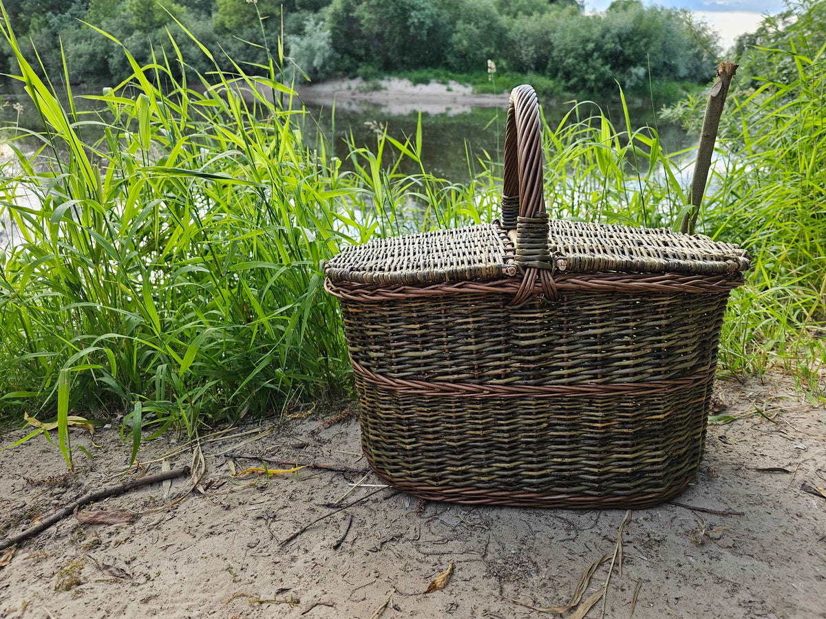 Uždaryta pikniko pintinė su rankena per vidurį ant smėlėto upės kranto tarp aukštos žolės